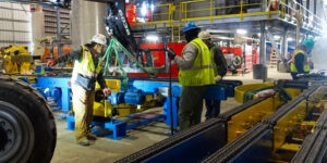 Workers securing blue and yellow machinery in a factory with industrial equipment.