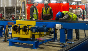 Workers discussing beside blue and yellow industrial machinery; one rests on the equipment.