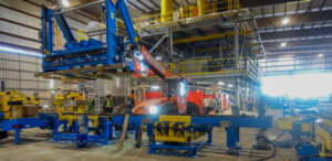 A forklift positions a large blue machine part above yellow industrial equipment in a factory.