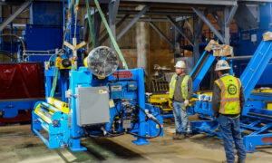 Workers guiding the installation of blue industrial machinery with yellow components in a factory.