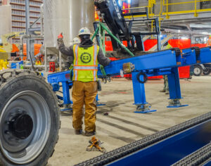 Worker in a safety vest guiding blue industrial equipment inside a factory.