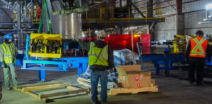 Workers in a factory installing yellow and blue industrial equipment on wooden pallets.