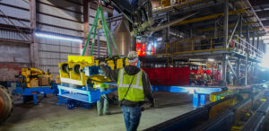 Worker guiding a suspended yellow machine part inside a factory with industrial equipment.