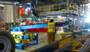 Workers positioning a suspended blue machine part inside an industrial factory.