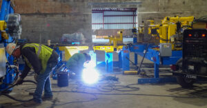 Worker welding near blue and yellow industrial machinery inside a factory.